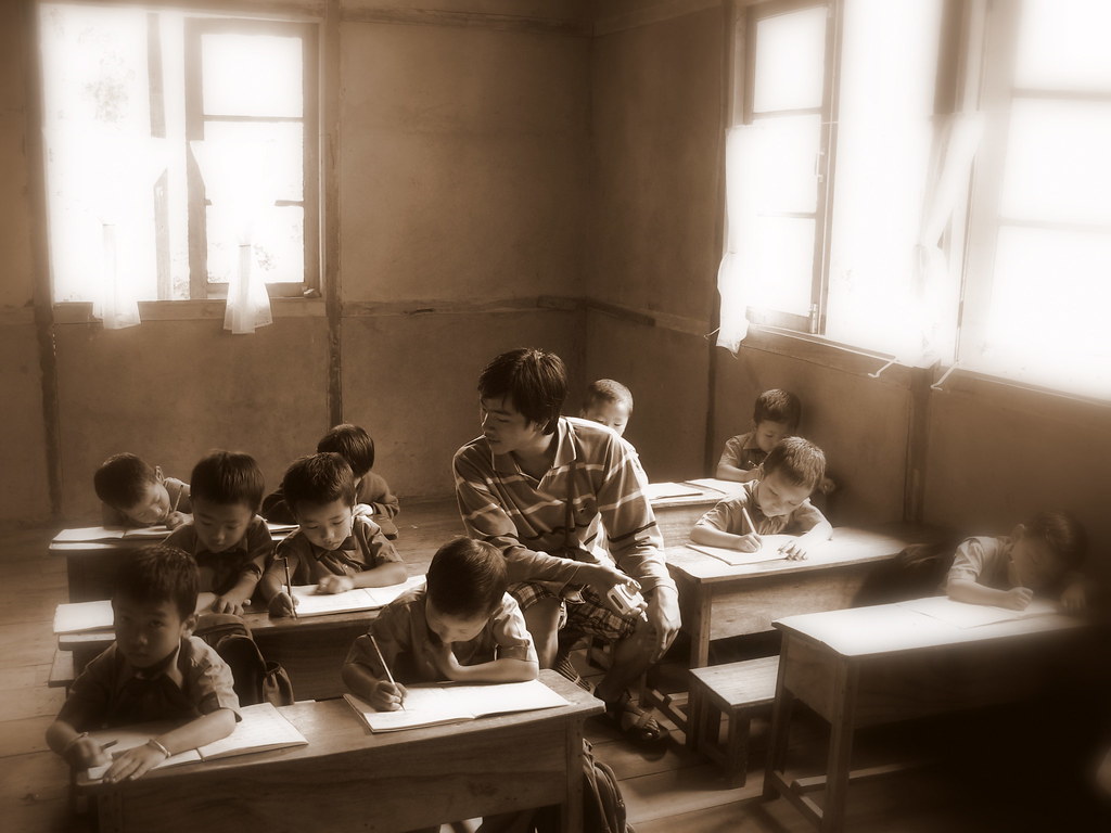 kids writing at desks in a classroom with a teacher checking their work