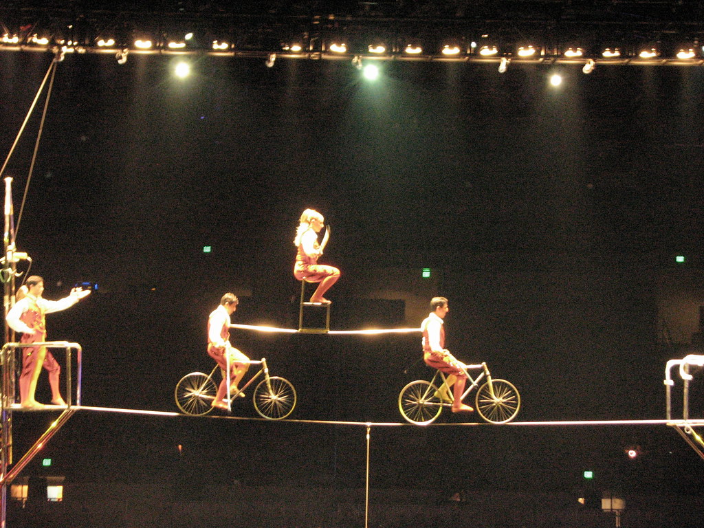 person on chair suspended between two cyclists riding across a tightrope