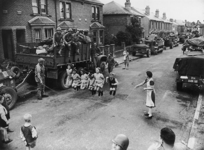 Wide shot of a street in which US soldiers help children play jump rope in World War 2