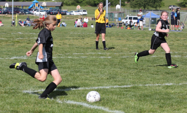 girls playing soccer on a grassy field passing the ball with referee in background